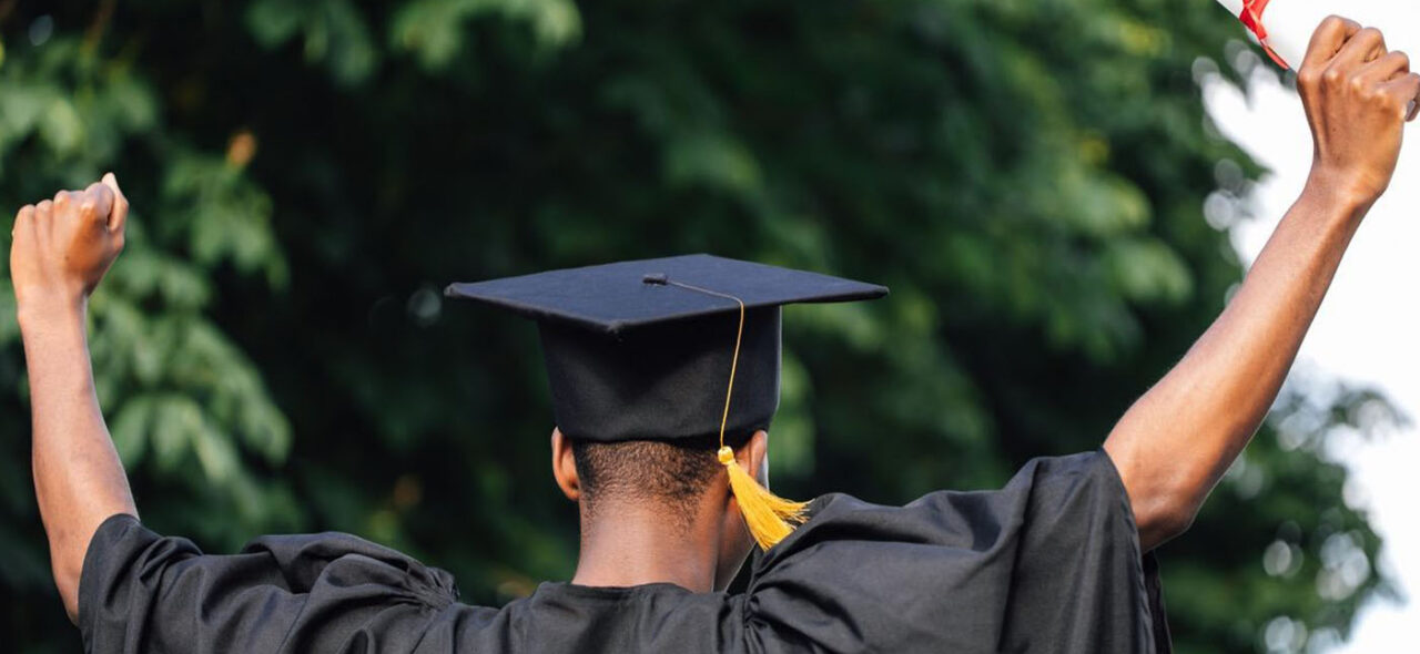 A student cheering in a graduation robe and hat, holding a diploma
